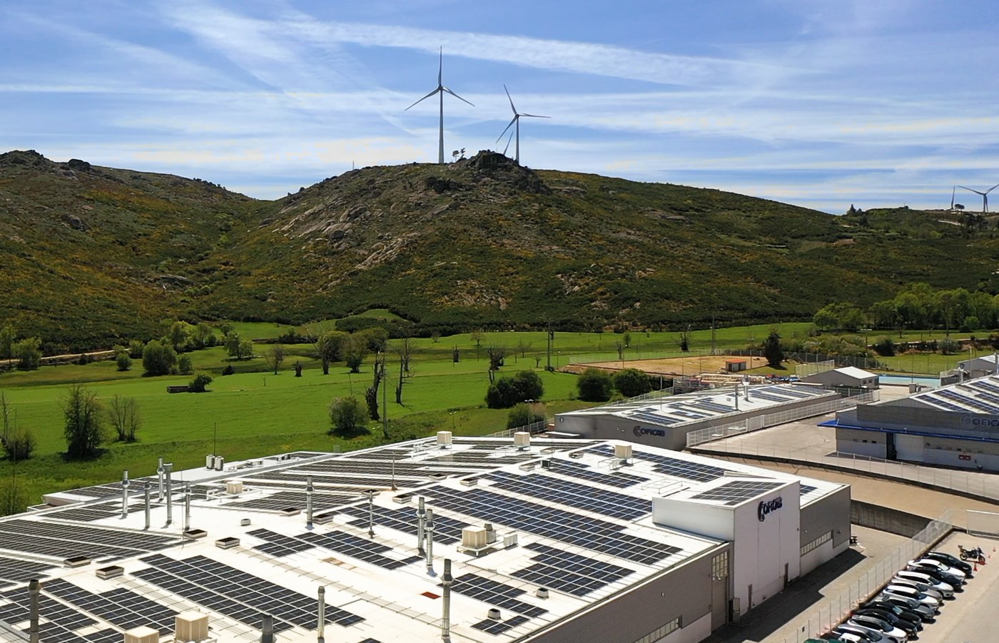 Fábrica da COFICAB com painéis solares na cobertura e turbinas eólicas na colina, ao fundo